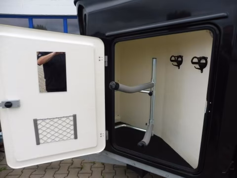 Offener schwarzer Kunststoffkasten mit weißer Tür, Metallleiter und Befestigungshaken im Inneren sichtbar.