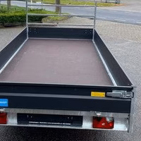 Schwarzer Kastenanhänger mit offener Ladefläche, Heckleuchten und Deichsel, geparkt auf grauem Untergrund.