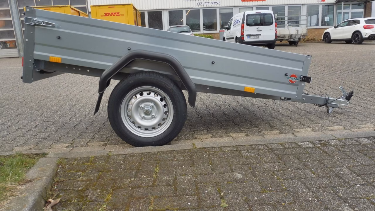 Grauer Anhänger mit offener Ladefläche auf Parkplatz, Seitenansicht mit Rad und Reifen sichtbar.