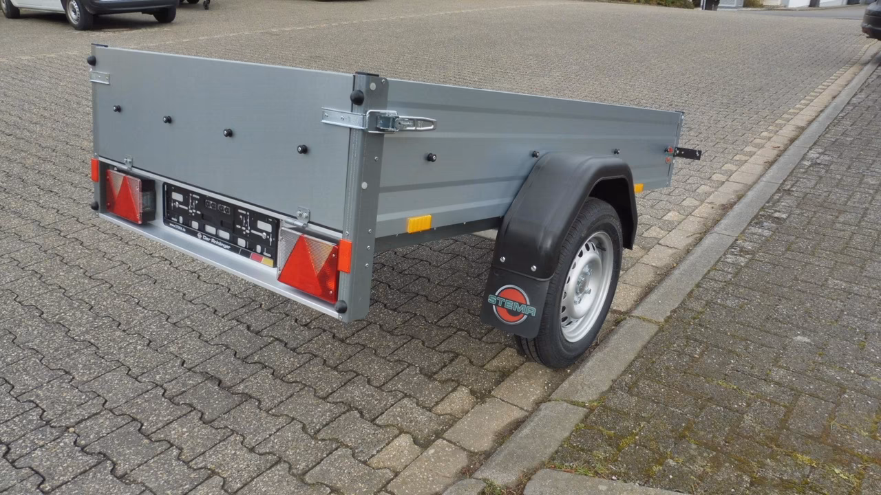 Grauer Anhänger mit rotem Rücklicht und schwarzem Rad auf gepflastertem Parkplatz geparkt.