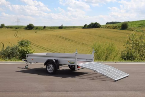 Silberner Anhänger mit Auffahrtsrampe auf Asphaltstraße vor grünem Feld und bewölktem Himmel.