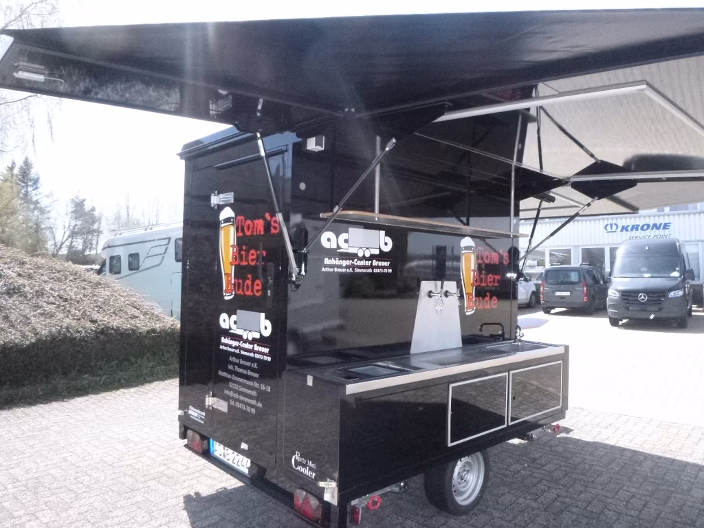 Schwarzer Imbisswagen mit ausgefahrenem Dach und Theke, geparkt auf Pflasterplatz vor Servicestelle.