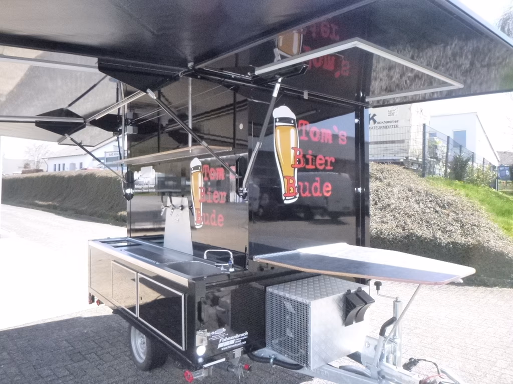 Mobiler Imbissstand mit schwarzem Dach, Grillbereich und Getränkespender auf Rädern.