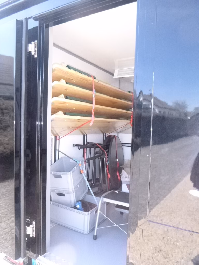 Geöffnete Transportbox mit Holzbalken, Fahrrad und weißen Behältern, gesichert mit roten Seilen.