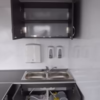 Kleine Küchenzeile mit Spüle, offenem Unterschrank mit Rohren und Behältern sowie Wandschrank darüber.