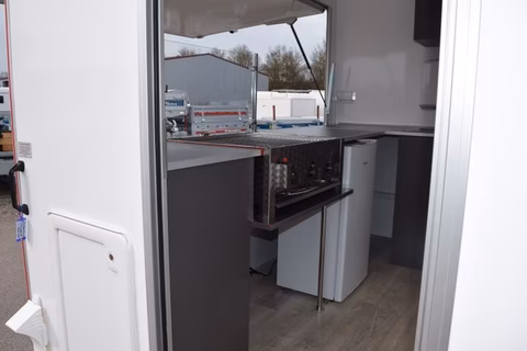 Innenansicht eines Wohnwagens mit Küche, Herd, Spüle und Kühlschrank, Fenster mit Außenblick.