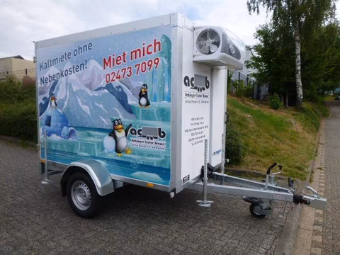 Kühlanhänger mit Pinguin-Motiv und Aufschrift "Kaltmiete ohne Nebenkosten! Miet mich 02473 7099" auf Parkplatz.