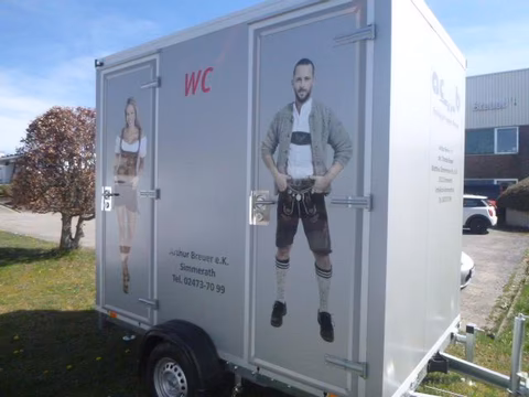 Mobiles WC-Häuschen mit Abbildungen einer Frau und eines Mannes in Tracht, geparkt auf einem Parkplatz.