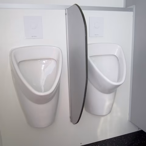 Zwei weiße Urinale an einer Wand mit Spülknöpfen und einem schwarzen Schlauch zwischen ihnen.