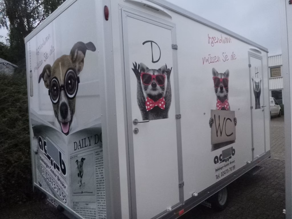 Weißer Wohnwagen mit bunten Tierillustrationen: Hund mit Brille, Waschbären mit Sonnenbrille und Fliege, WC-Schild.