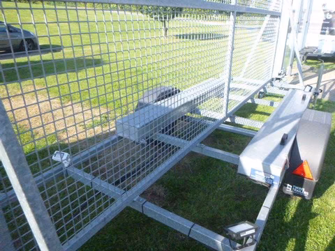 Anhänger mit Gitterbox und offener Rampe auf grüner Wiese, Seitenansicht.