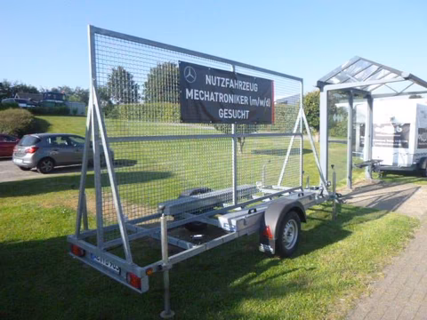 Anhänger mit Netzgitter und Schild "Nutzfahrzeug Mechatroniker gesucht" auf grüner Wiese.